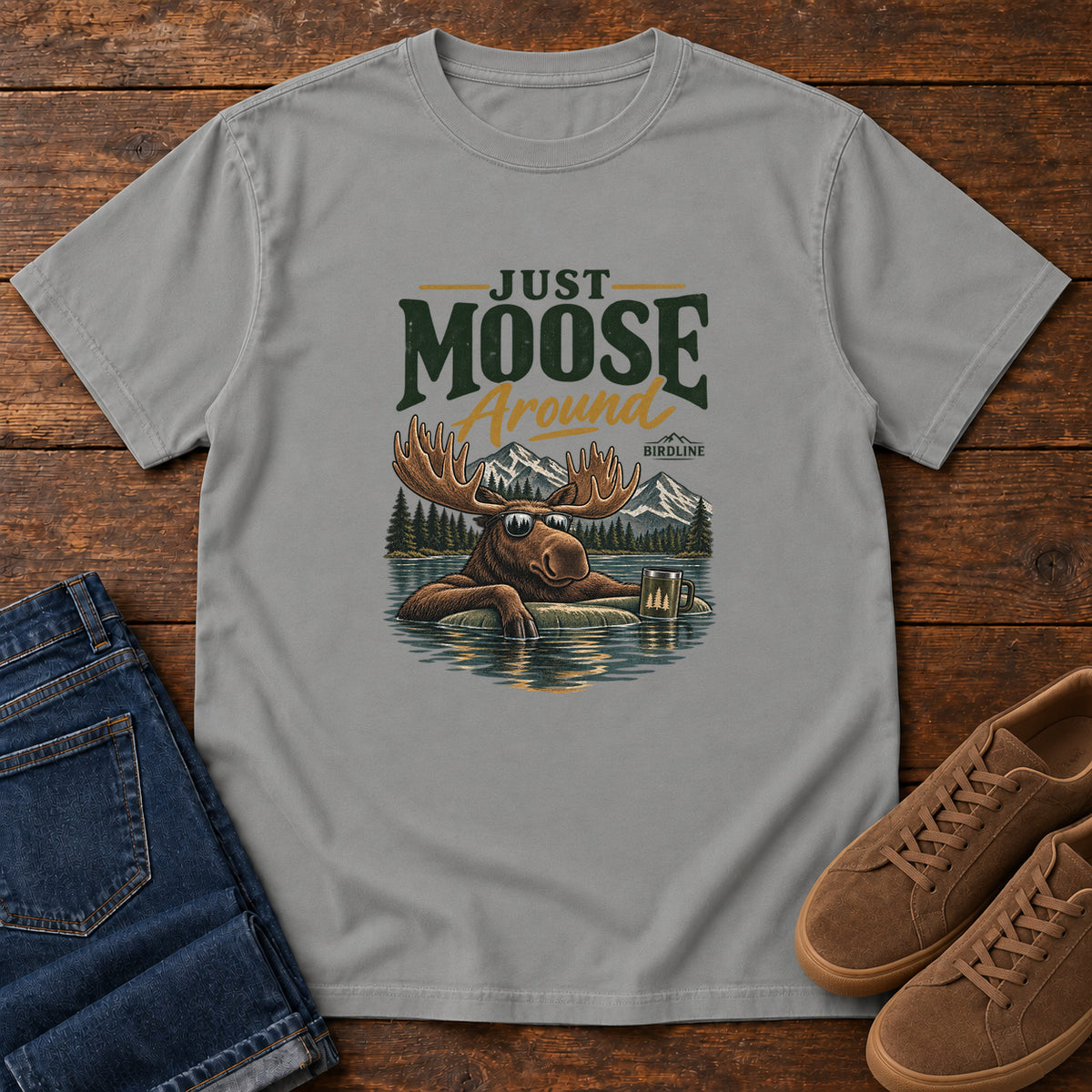 Grey Just Moose Around T-Shirt with a moose wearing sunglasses in a lake beneath mountains and slogan lettering; front flat lay on wood.
