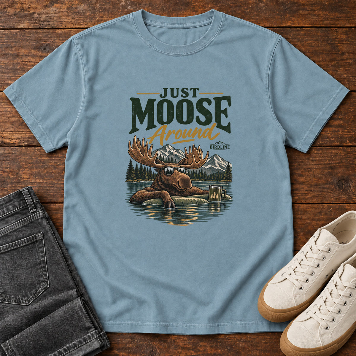 Ice Blue Just Moose Around T-Shirt with a moose wearing sunglasses in a lake beneath mountains and slogan lettering; front flat lay on wood.