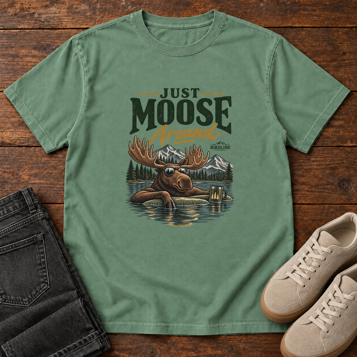 Light Green Just Moose Around T-Shirt with a moose wearing sunglasses in a lake beneath mountains and slogan lettering; front flat lay on wood.