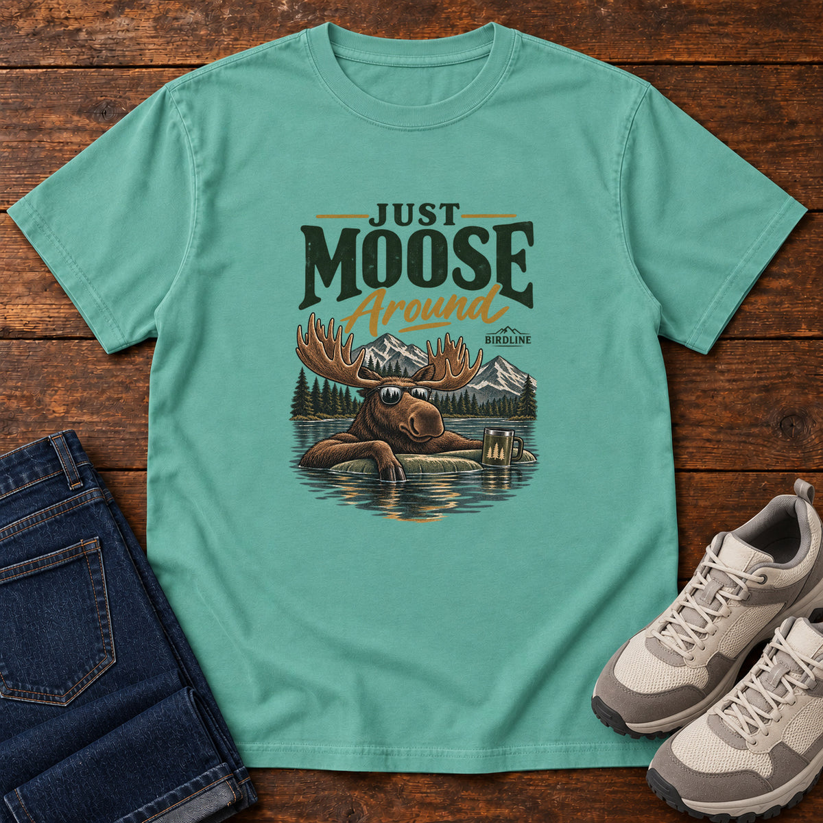 Seafoam Just Moose Around T-Shirt with a moose wearing sunglasses in a lake beneath mountains and slogan lettering; front flat lay on wood.
