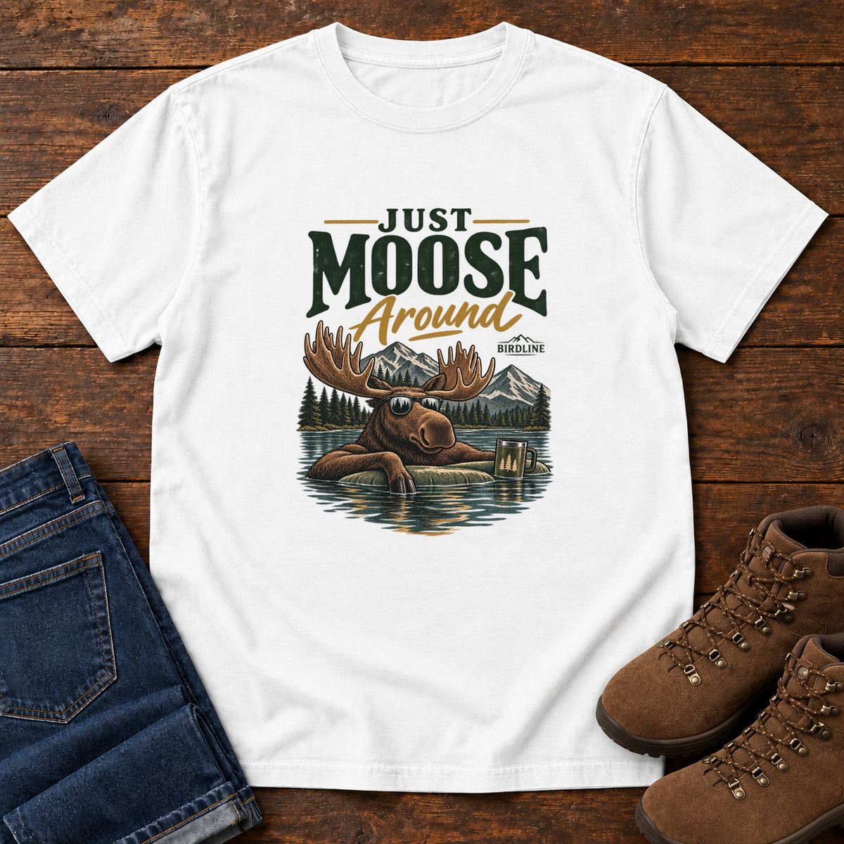 White Just Moose Around T-Shirt with a moose wearing sunglasses in a lake beneath mountains and slogan lettering; front flat lay on wood.