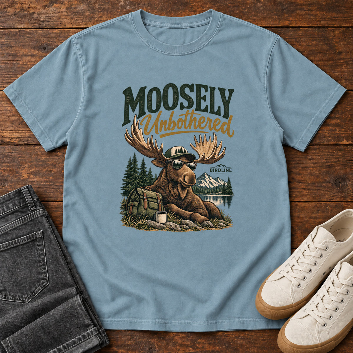 Ice Blue Moosely Unbothered T-Shirt with a lounging moose in sunglasses beside a backpack and mug, front flat lay on wood.