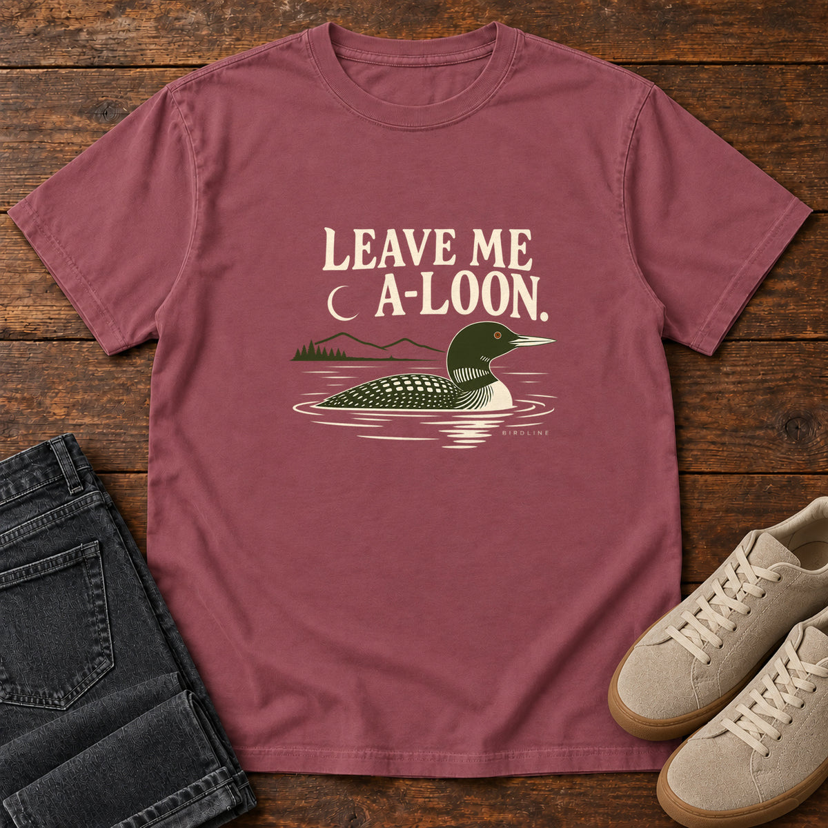 Brick Leave Me A Loon T-Shirt with a green-and-cream waterbird floating below a crescent moon and cream lettering, front flat lay on wood.