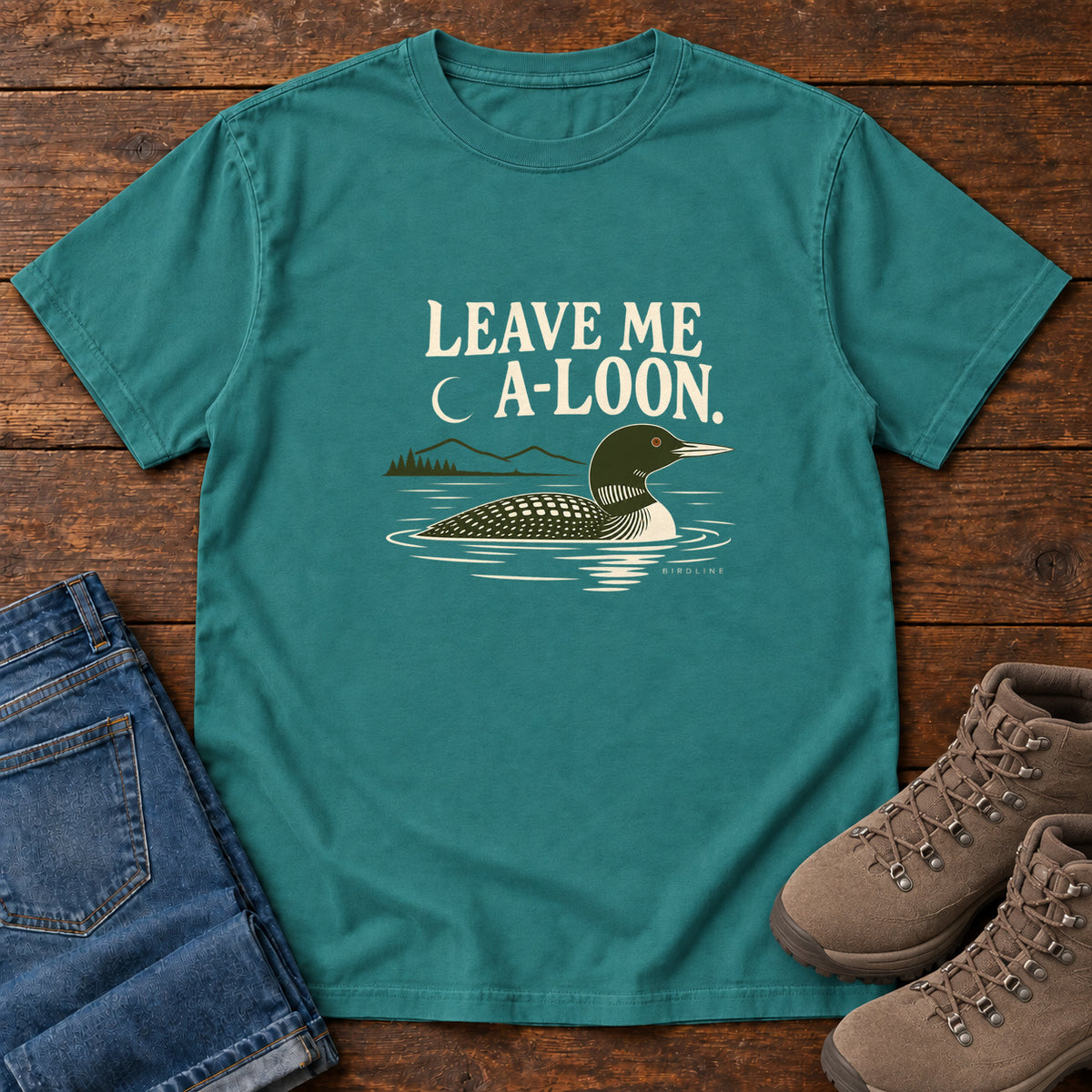 Emerald Leave Me A Loon T-Shirt with a green-and-cream waterbird floating below a crescent moon and cream lettering, front flat lay on wood.