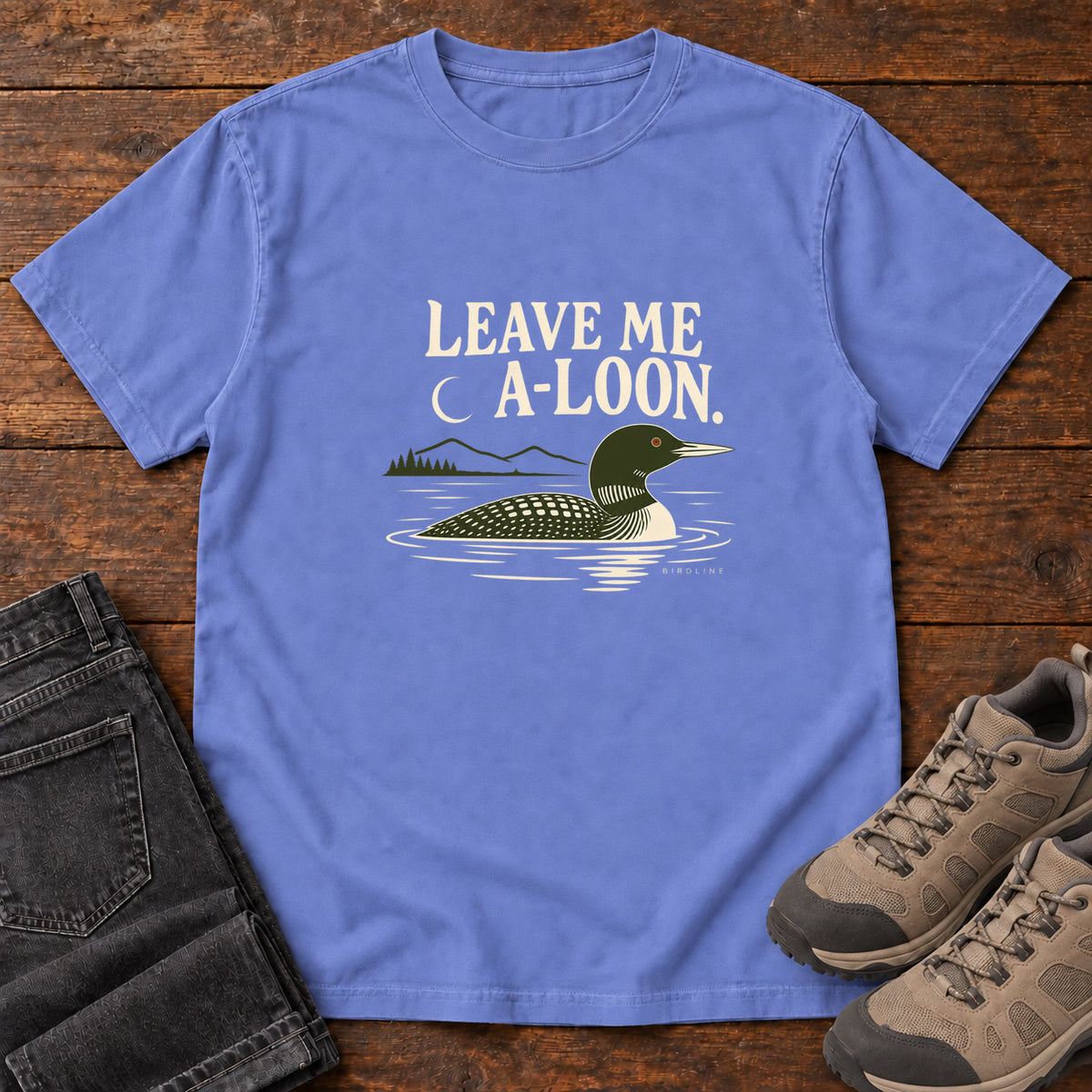 Flo Blue Leave Me A Loon T-Shirt with a green-and-cream waterbird floating below a crescent moon and cream lettering, front flat lay on wood.