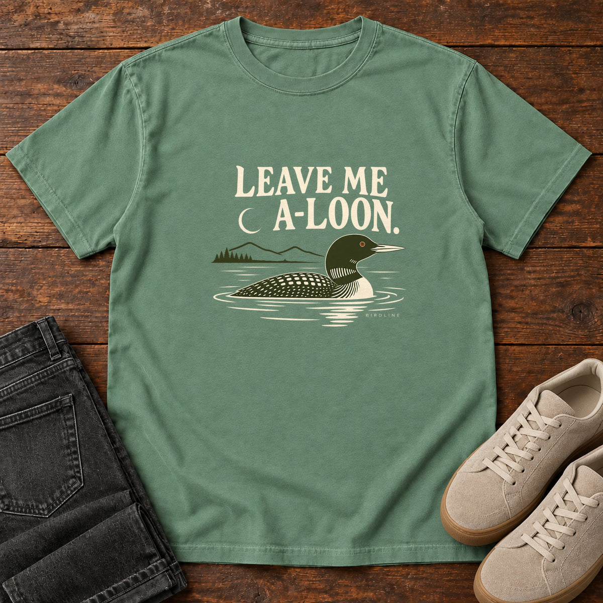 Light Green Leave Me A Loon T-Shirt with a green-and-cream waterbird floating below a crescent moon and cream lettering, front flat lay on wood.