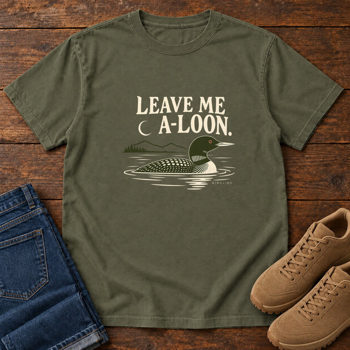 Sage Leave Me A Loon T-Shirt with a green-and-cream waterbird floating below a crescent moon and cream lettering, front flat lay on wood.