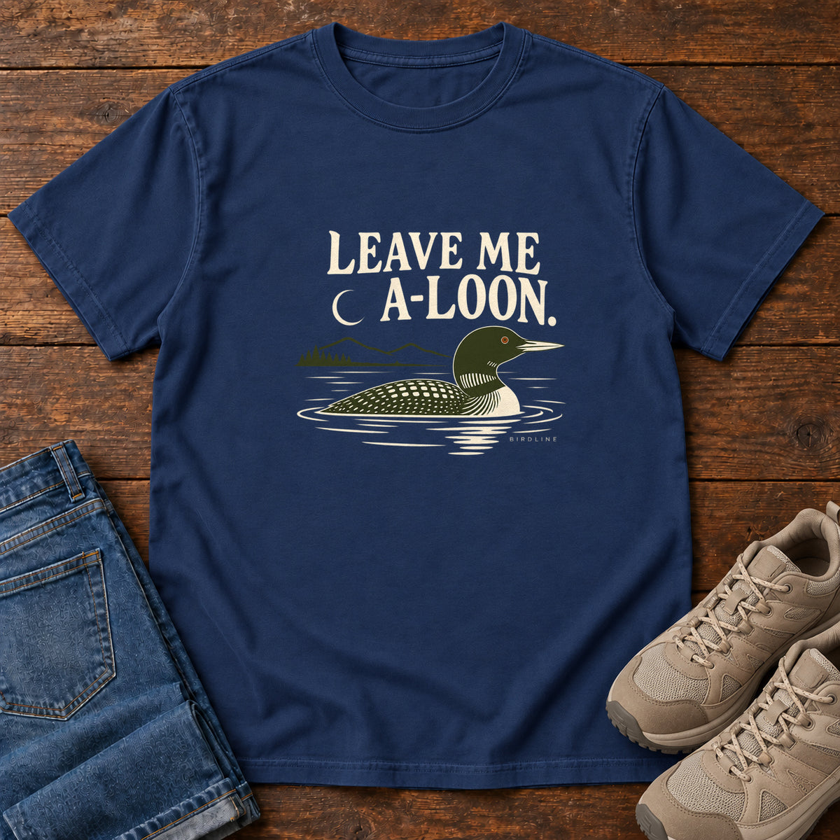 True Navy Leave Me A Loon T-Shirt with a green-and-cream waterbird floating below a crescent moon and cream lettering, front flat lay on wood.