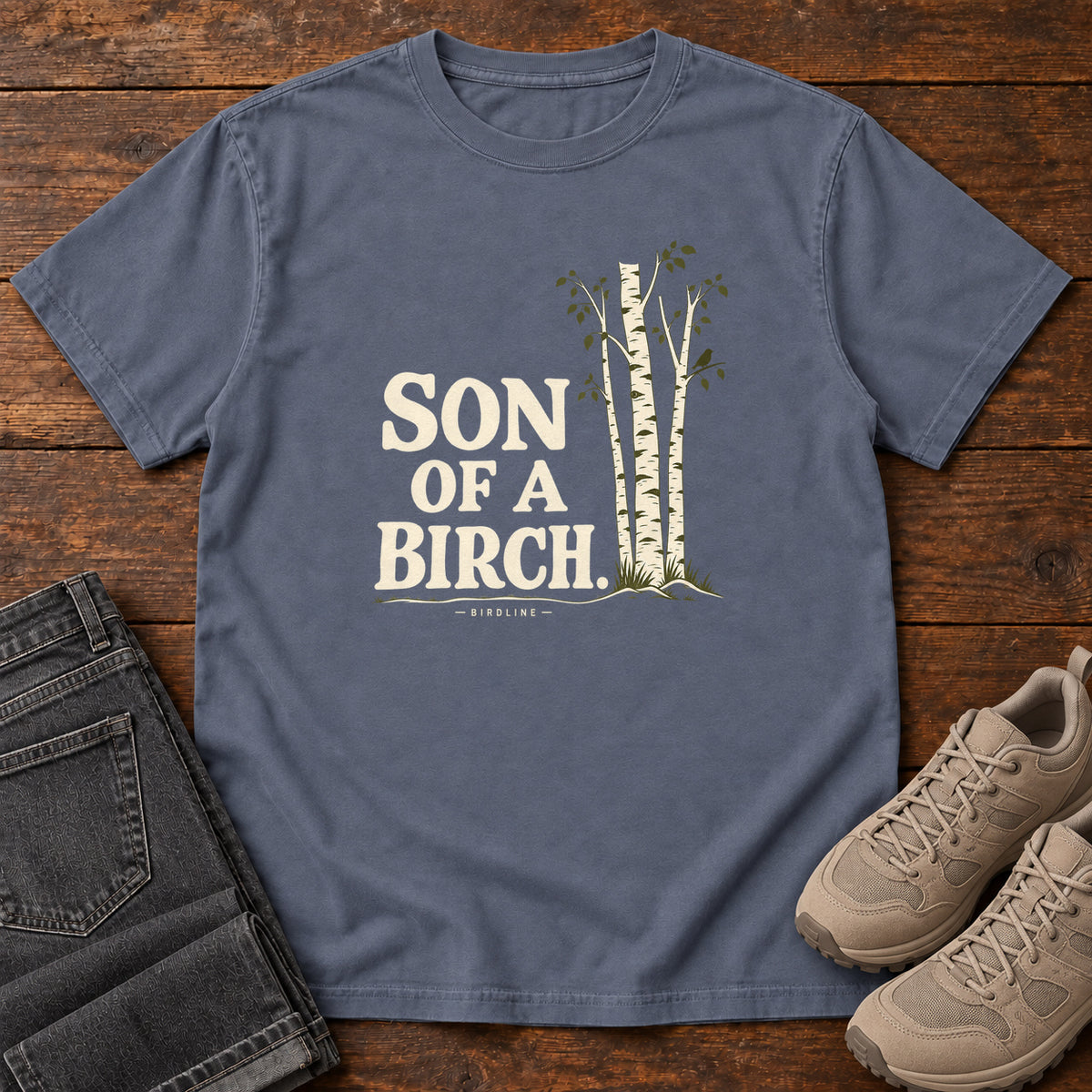 Denim Son of a Birch T-Shirt with cream lettering beside slender white-barked trees, front flat lay on wood.