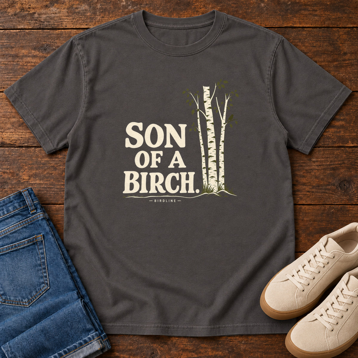 Graphite Son of a Birch T-Shirt with cream lettering beside slender white-barked trees, front flat lay on wood.