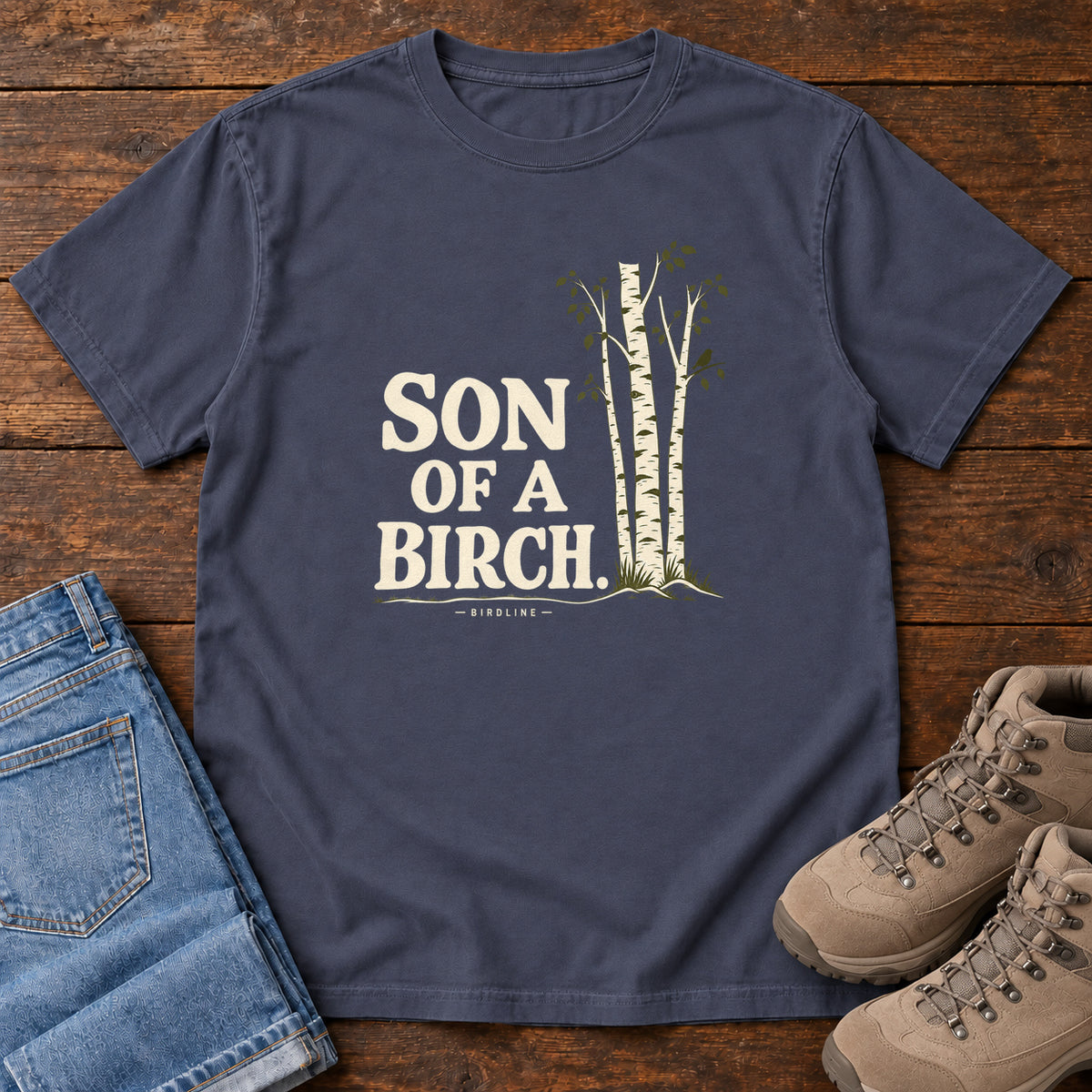 Navy Son of a Birch T-Shirt with cream lettering beside slender white-barked trees, front flat lay on wood.