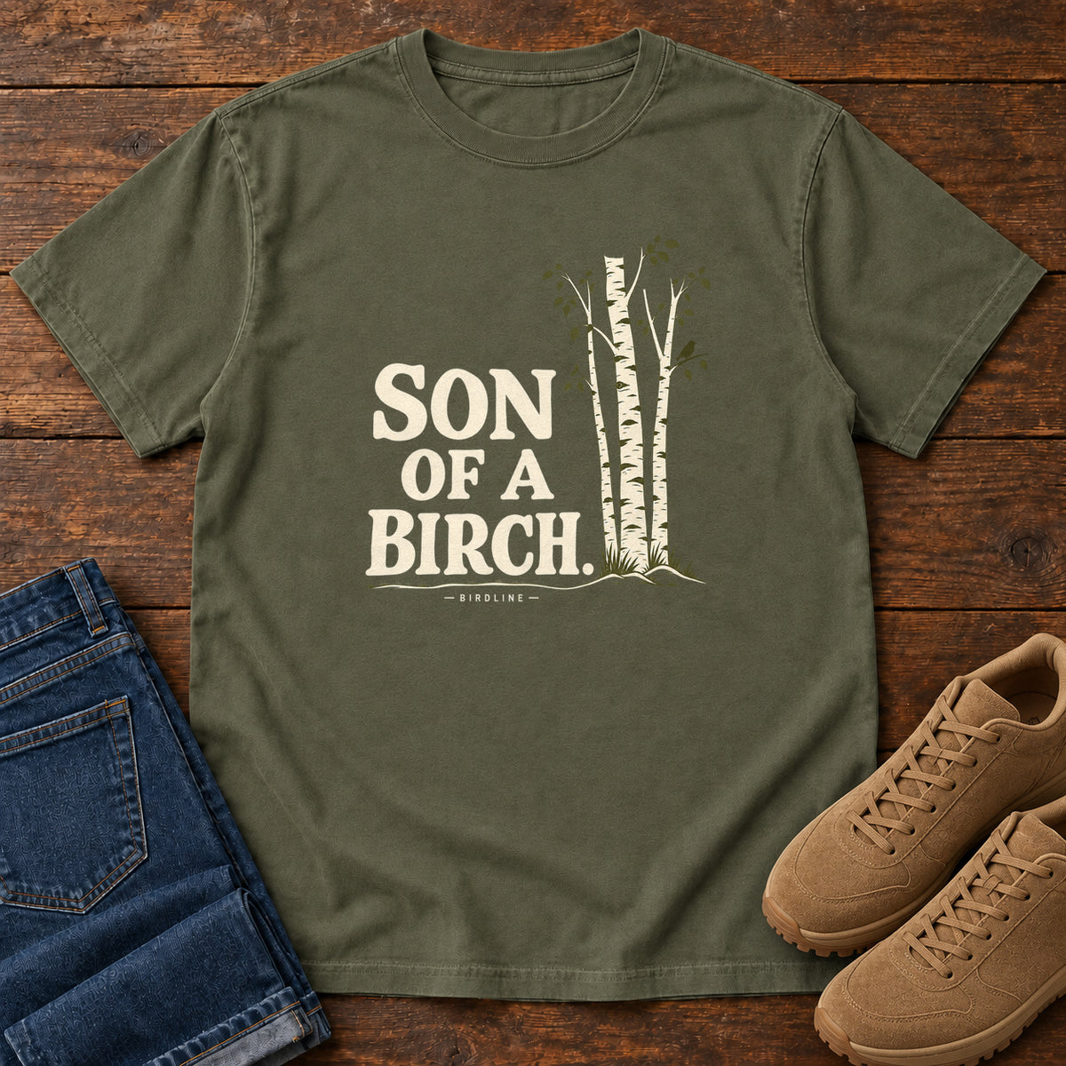 Sage Son of a Birch T-Shirt with cream lettering beside slender white-barked trees, front flat lay on wood.
