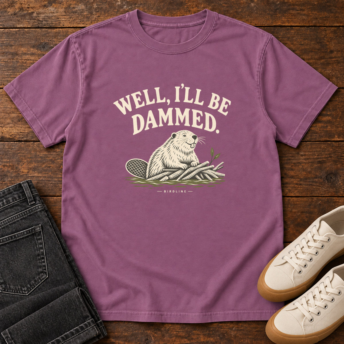 Well I'll Be Dammed T-Shirt in Berry, front laid flat on wood, featuring a cream beaver sitting beside branches beneath curved slogan lettering.