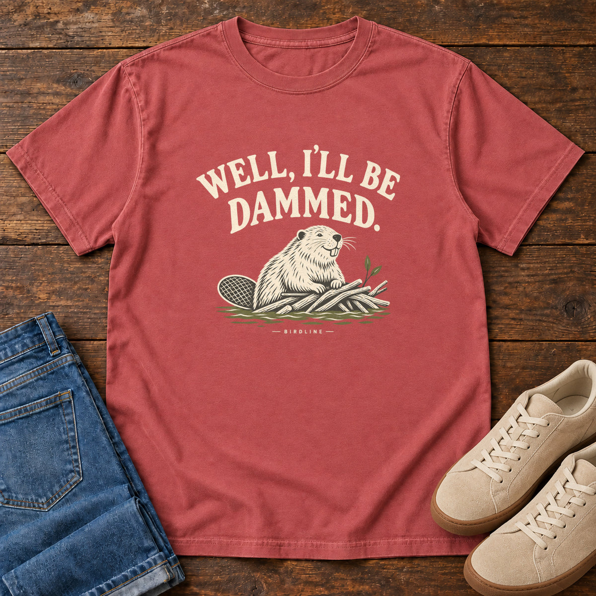 Well I'll Be Dammed T-Shirt in Crimson, front laid flat on wood, featuring a cream beaver sitting beside branches beneath curved slogan lettering.