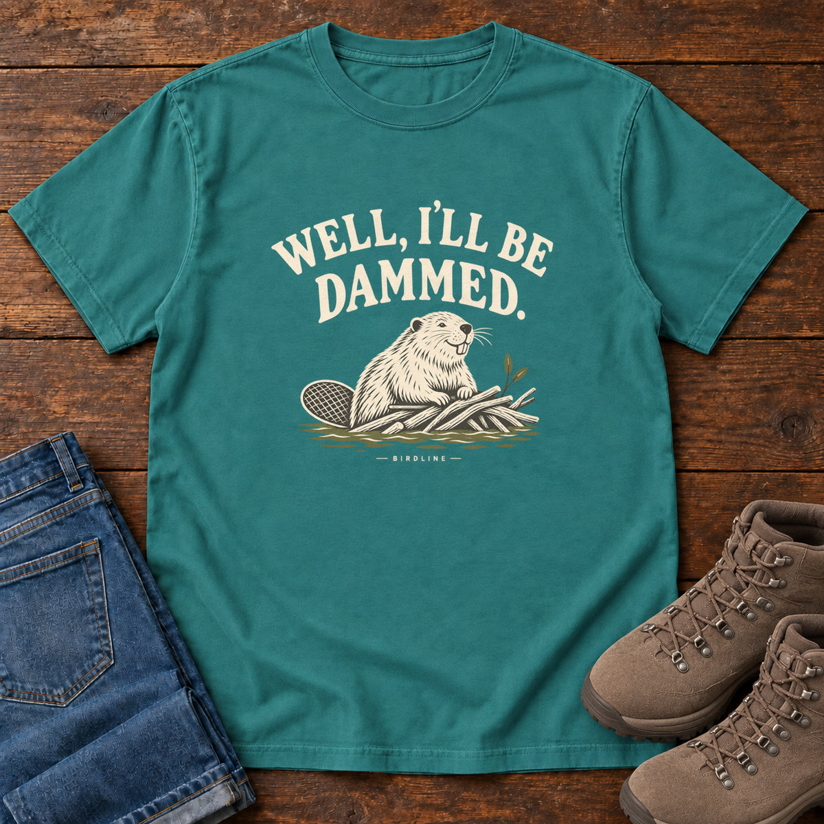Well I'll Be Dammed T-Shirt in Emerald, front laid flat on wood, featuring a cream beaver sitting beside branches beneath curved slogan lettering.