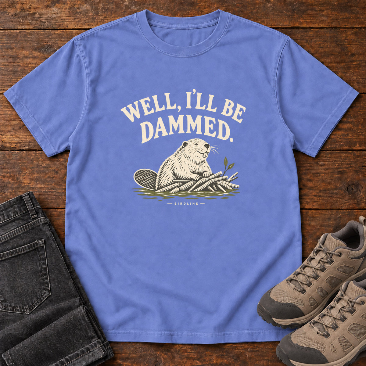 Well I'll Be Dammed T-Shirt in Flo Blue, front laid flat on wood, featuring a cream beaver sitting beside branches beneath curved slogan lettering.