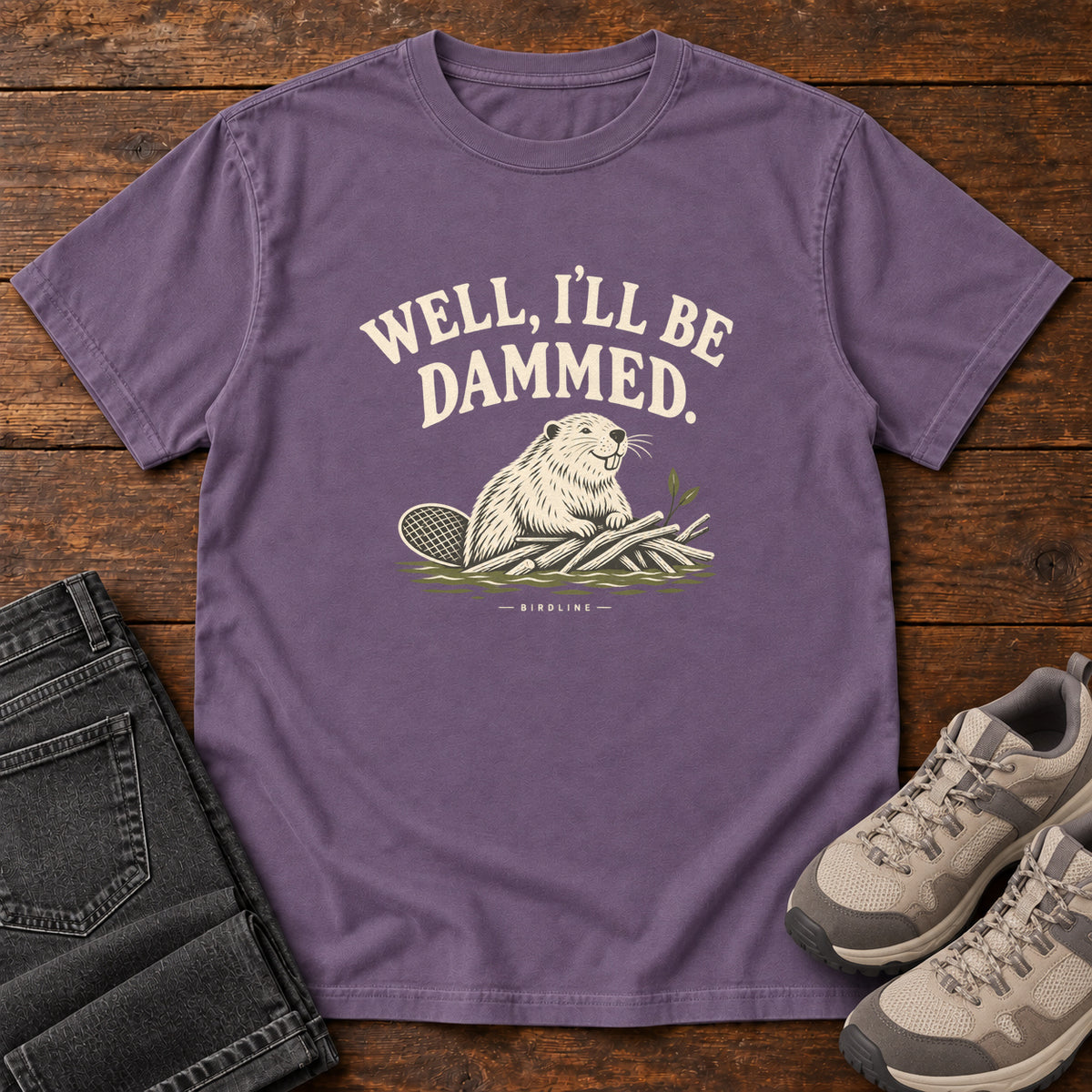 Well I'll Be Dammed T-Shirt in Grape, front laid flat on wood, featuring a cream beaver sitting beside branches beneath curved slogan lettering.