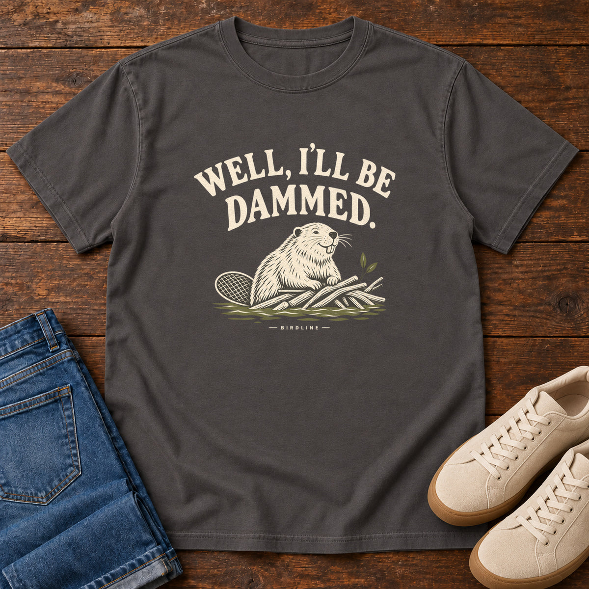 Well I'll Be Dammed T-Shirt in Graphite, front laid flat on wood, featuring a cream beaver sitting beside branches beneath curved slogan lettering.