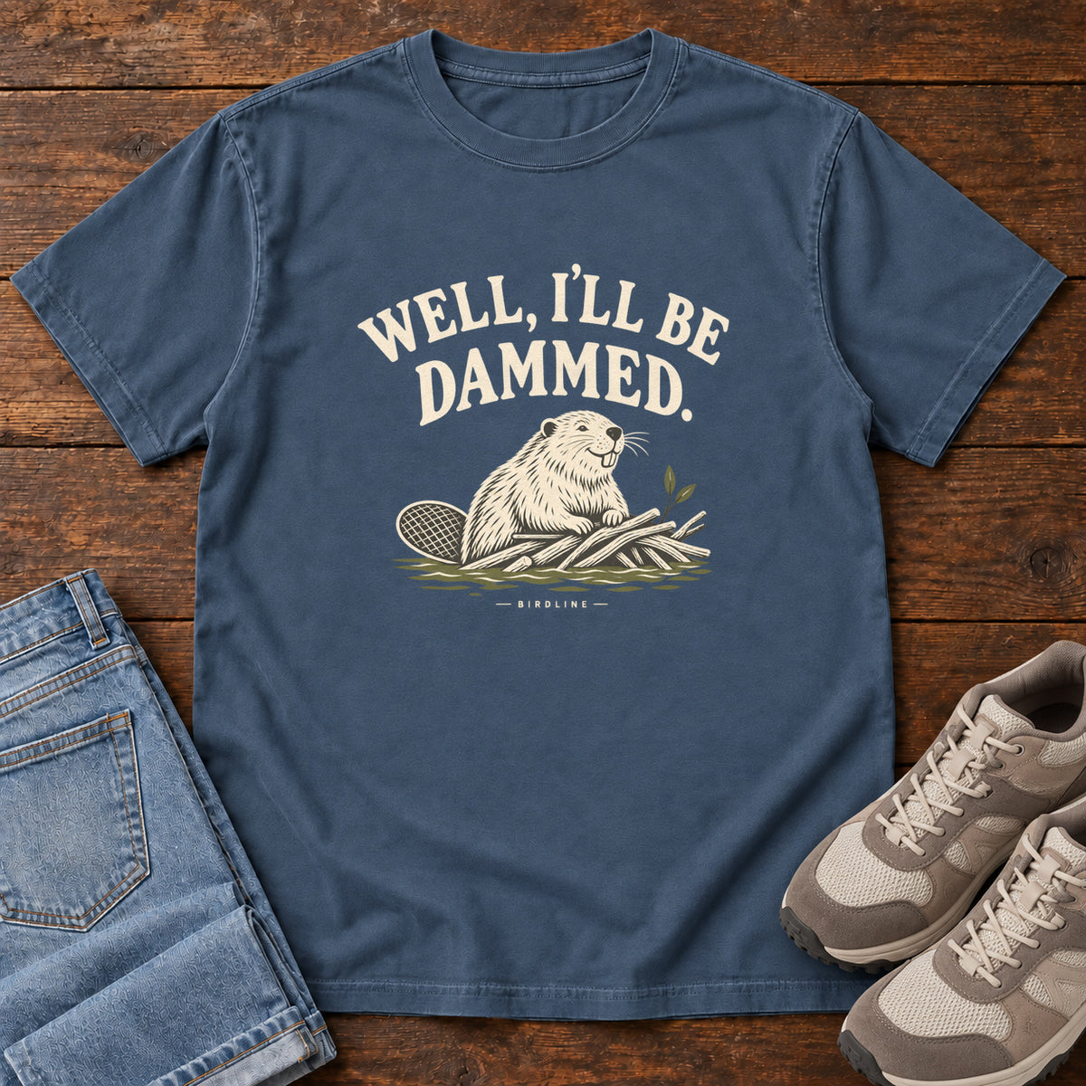 Well I'll Be Dammed T-Shirt in Midnight, front laid flat on wood, featuring a cream beaver sitting beside branches beneath curved slogan lettering.