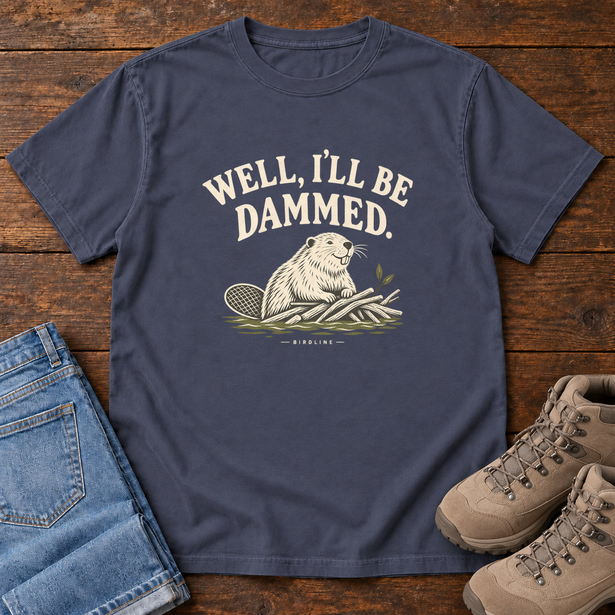 Well I'll Be Dammed T-Shirt in Navy, front laid flat on wood, featuring a cream beaver sitting beside branches beneath curved slogan lettering.