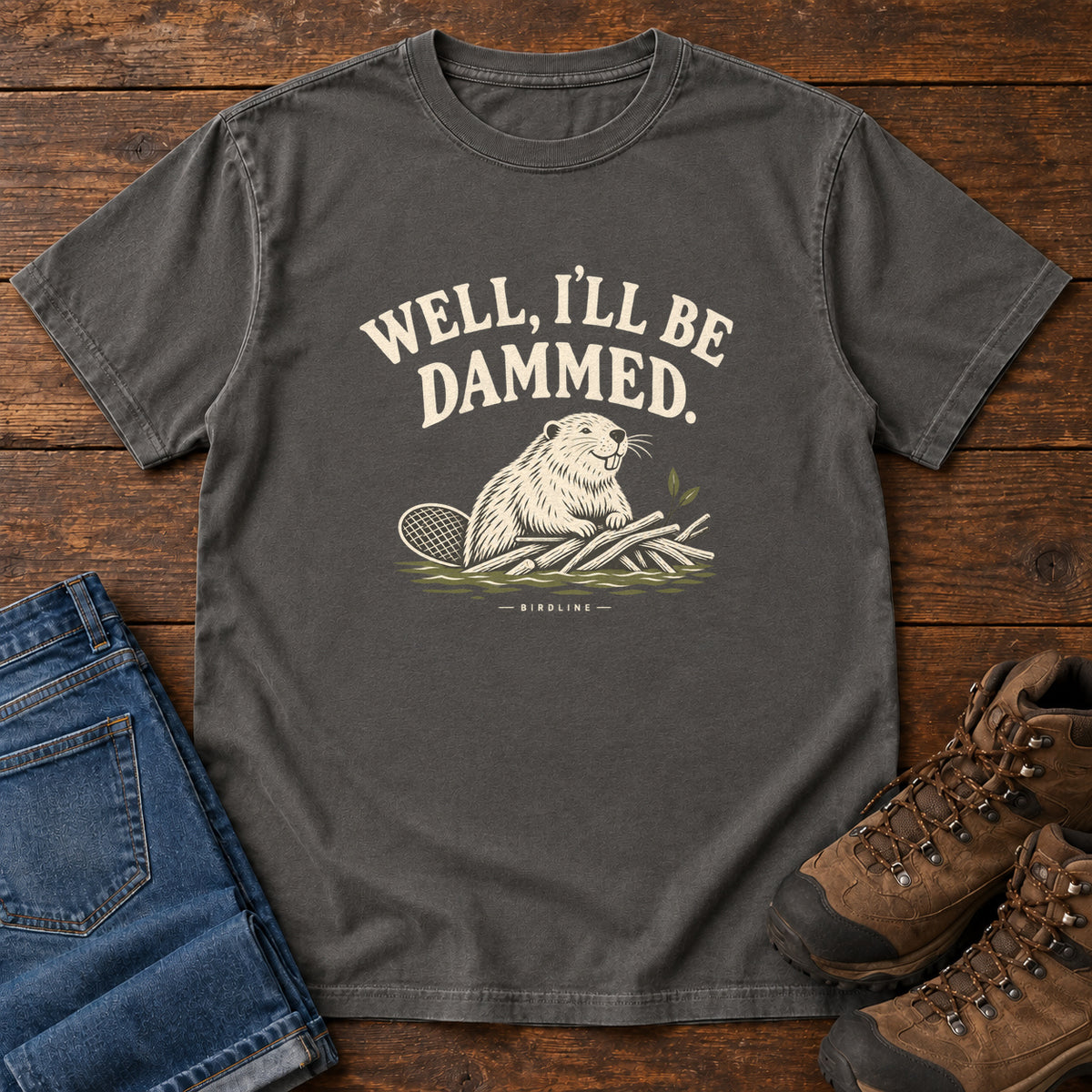 Well I'll Be Dammed T-Shirt in Pepper, front laid flat on wood, featuring a cream beaver sitting beside branches beneath curved slogan lettering.