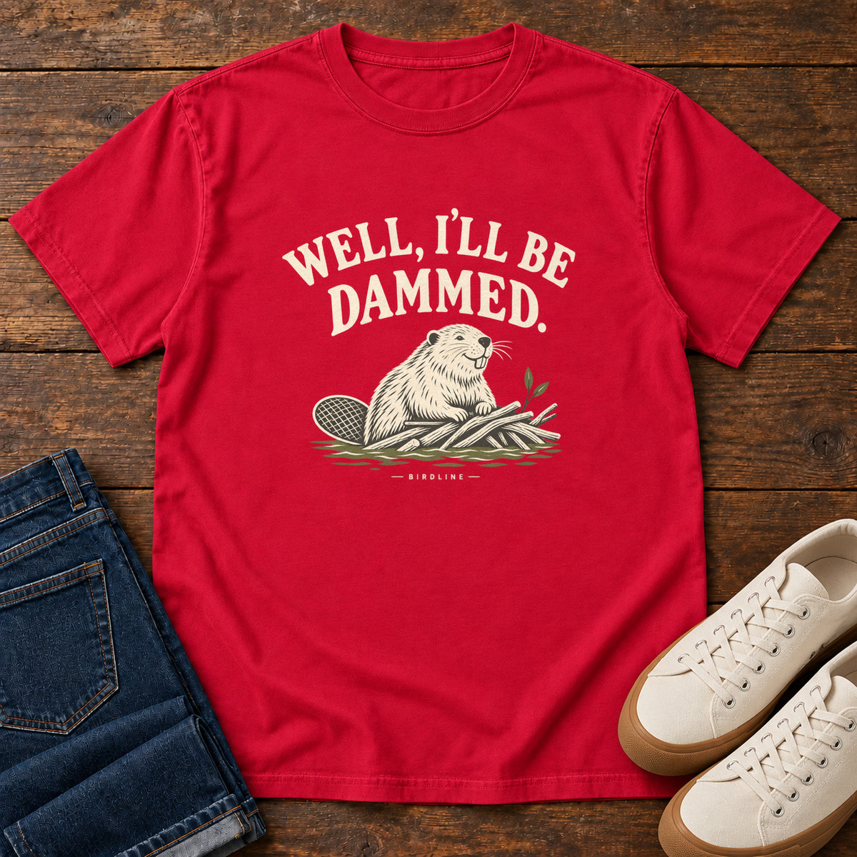 Well I'll Be Dammed T-Shirt in Red, front laid flat on wood, featuring a cream beaver sitting beside branches beneath curved slogan lettering.