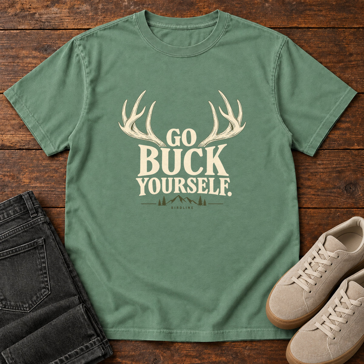 Light Green Go Buck Yourself T-Shirt with cream deer antlers framing Go Buck Yourself lettering, front flat lay on wood.