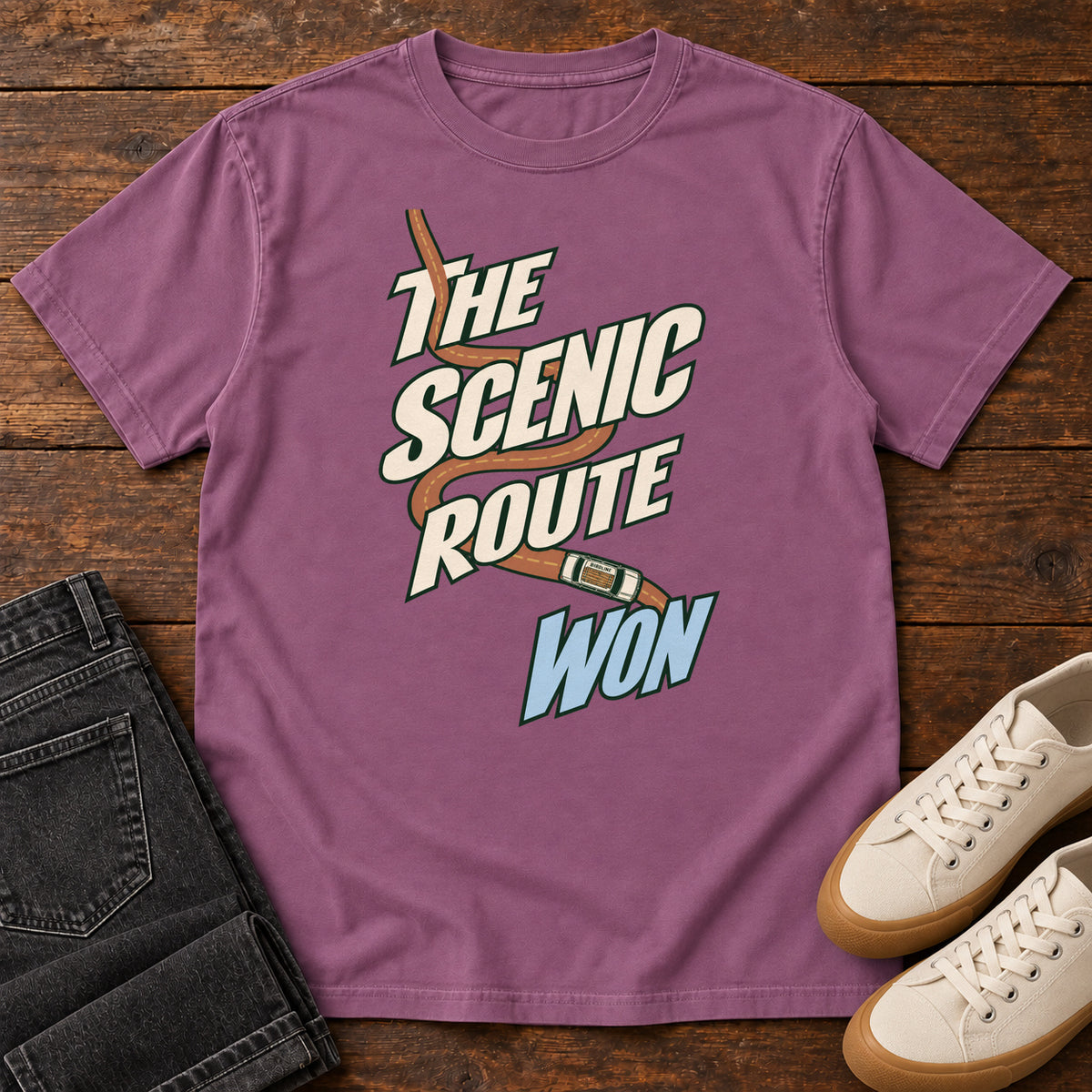 The Scenic Route Won T-Shirt in Berry, front laid flat on wood, featuring a winding orange road and tiny car behind slanted cream-and-blue slogan lettering.