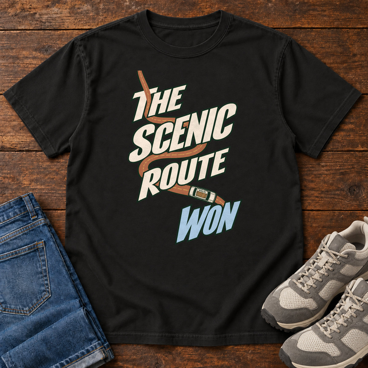 The Scenic Route Won T-Shirt in Black, front laid flat on wood, featuring a winding orange road and tiny car behind slanted cream-and-blue slogan lettering.