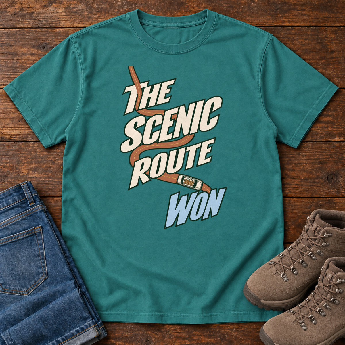 The Scenic Route Won T-Shirt in Emerald, front laid flat on wood, featuring a winding orange road and tiny car behind slanted cream-and-blue slogan lettering.