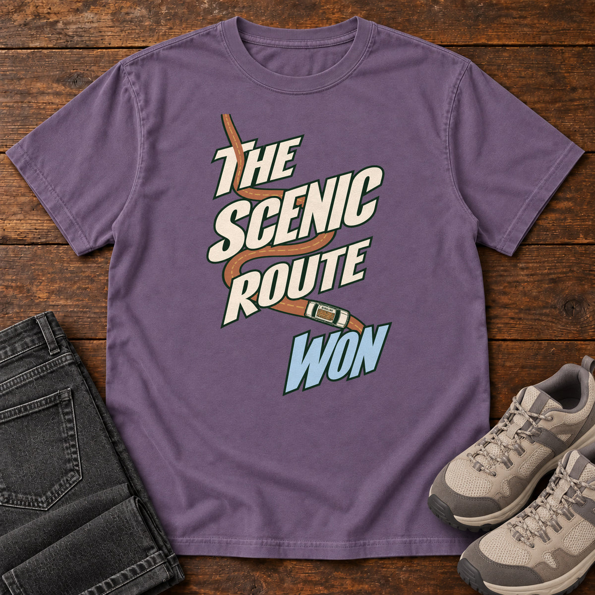 The Scenic Route Won T-Shirt in Grape, front laid flat on wood, featuring a winding orange road and tiny car behind slanted cream-and-blue slogan lettering.