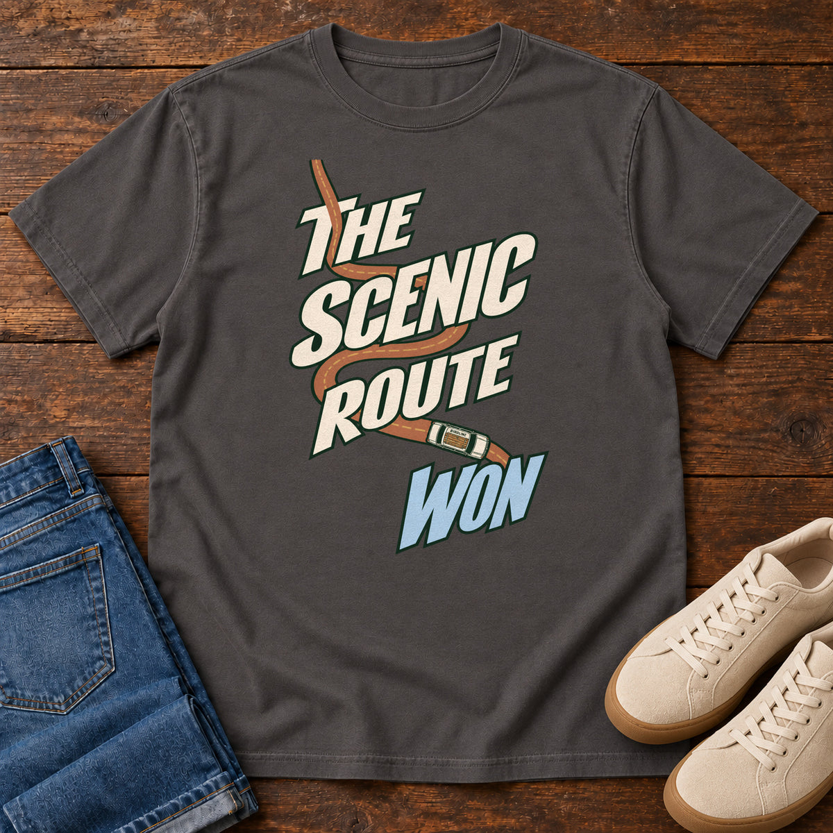 The Scenic Route Won T-Shirt in Graphite, front laid flat on wood, featuring a winding orange road and tiny car behind slanted cream-and-blue slogan lettering.