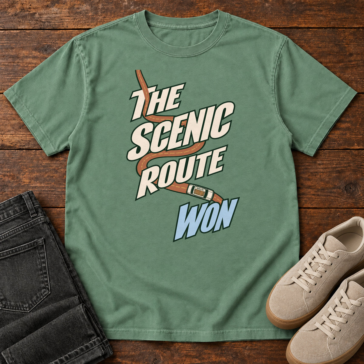 The Scenic Route Won T-Shirt in Light Green, front laid flat on wood, featuring a winding orange road and tiny car behind slanted cream-and-blue slogan lettering.