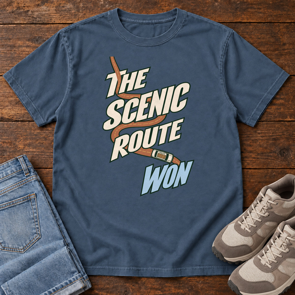 The Scenic Route Won T-Shirt in Midnight, front laid flat on wood, featuring a winding orange road and tiny car behind slanted cream-and-blue slogan lettering.