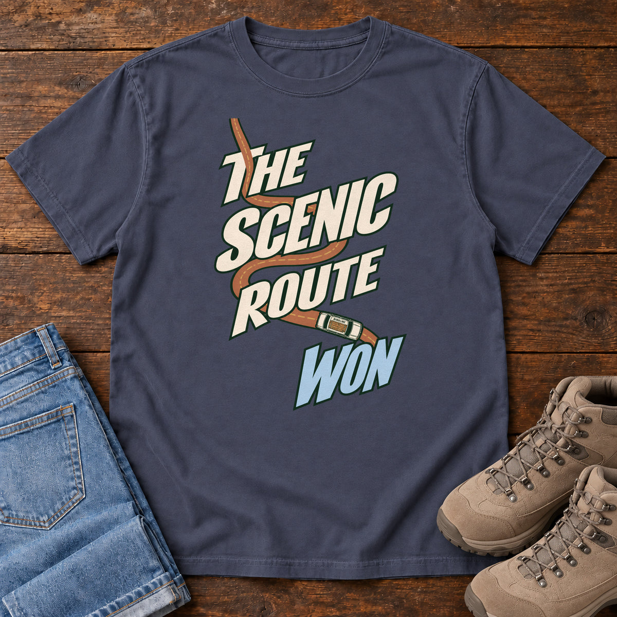 The Scenic Route Won T-Shirt in Navy, front laid flat on wood, featuring a winding orange road and tiny car behind slanted cream-and-blue slogan lettering.