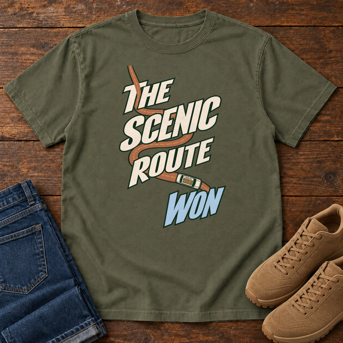 The Scenic Route Won T-Shirt in Sage, front laid flat on wood, featuring a winding orange road and tiny car behind slanted cream-and-blue slogan lettering.