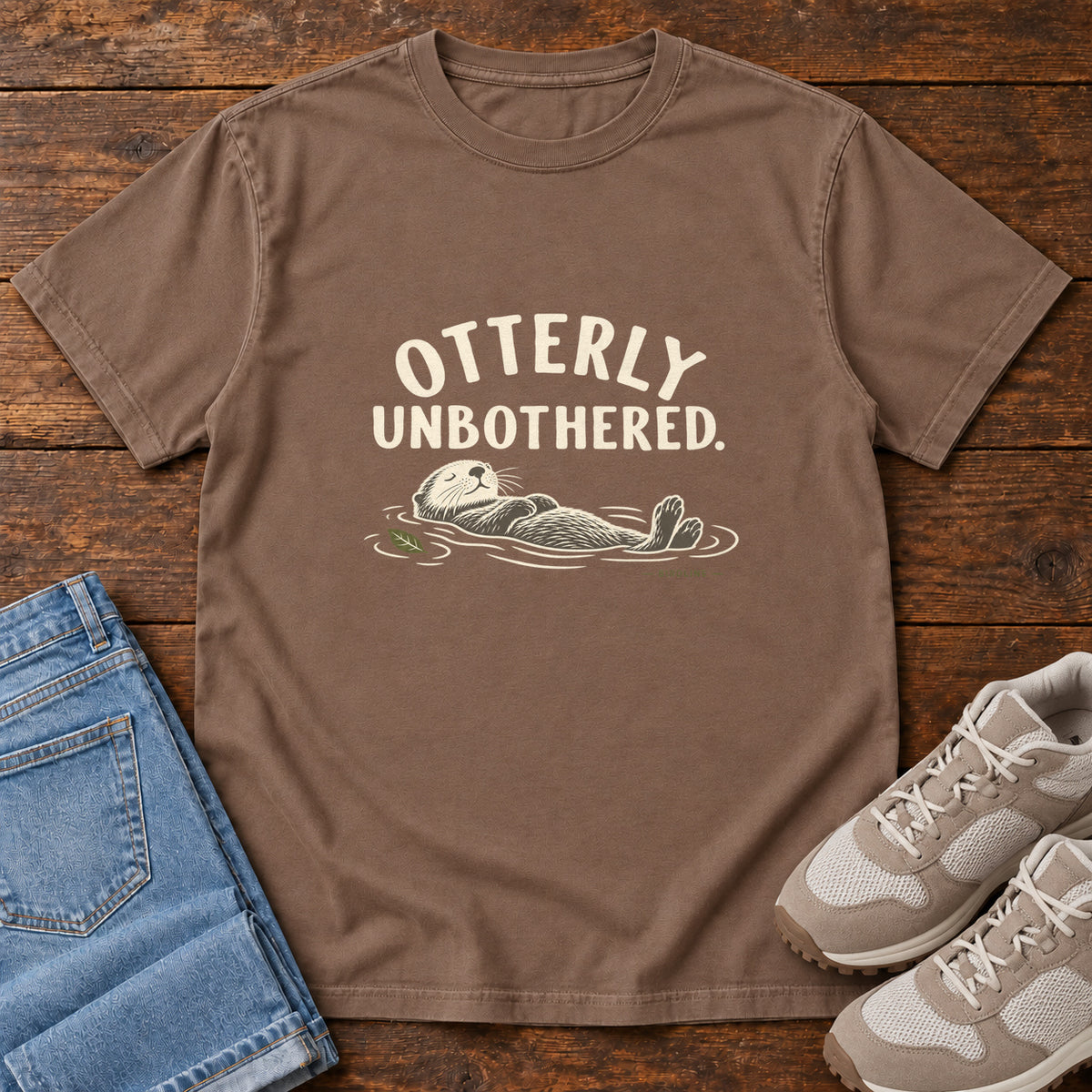 Espresso Otterly Unbothered T-Shirt with a cream line drawing of an otter floating on its back beneath curved lettering; front flat lay on wood.