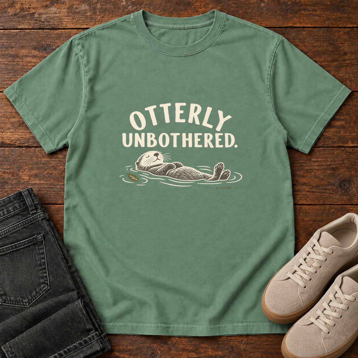 Light Green Otterly Unbothered T-Shirt with a cream line drawing of an otter floating on its back beneath curved lettering; front flat lay on wood.