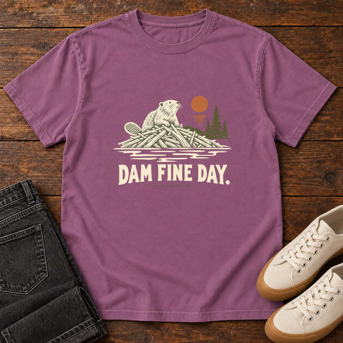 Berry Dam Fine Day T-Shirt with a beaver on a pile of branches beside evergreens and an orange sun; front flat lay on wood.