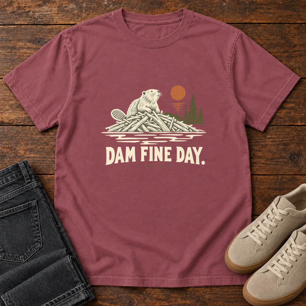 Brick Dam Fine Day T-Shirt with a beaver on a pile of branches beside evergreens and an orange sun; front flat lay on wood.