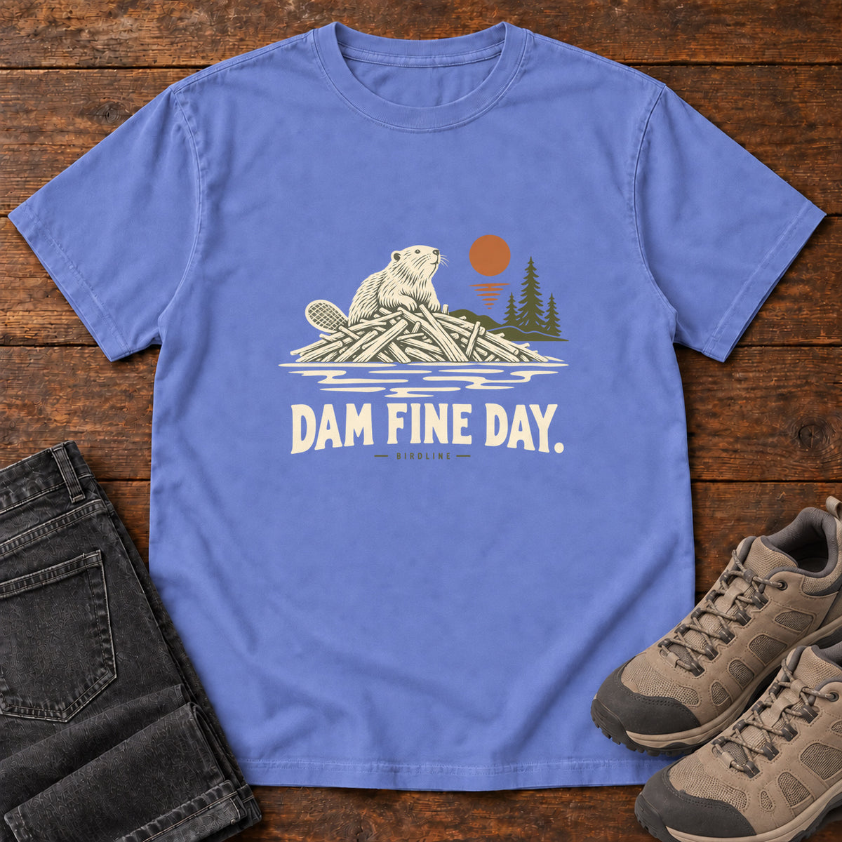 Flo Blue Dam Fine Day T-Shirt with a beaver on a pile of branches beside evergreens and an orange sun; front flat lay on wood.