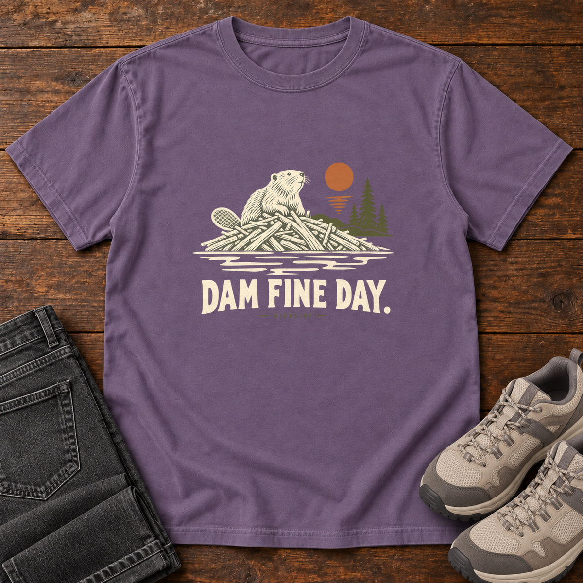 Grape Dam Fine Day T-Shirt with a beaver on a pile of branches beside evergreens and an orange sun; front flat lay on wood.