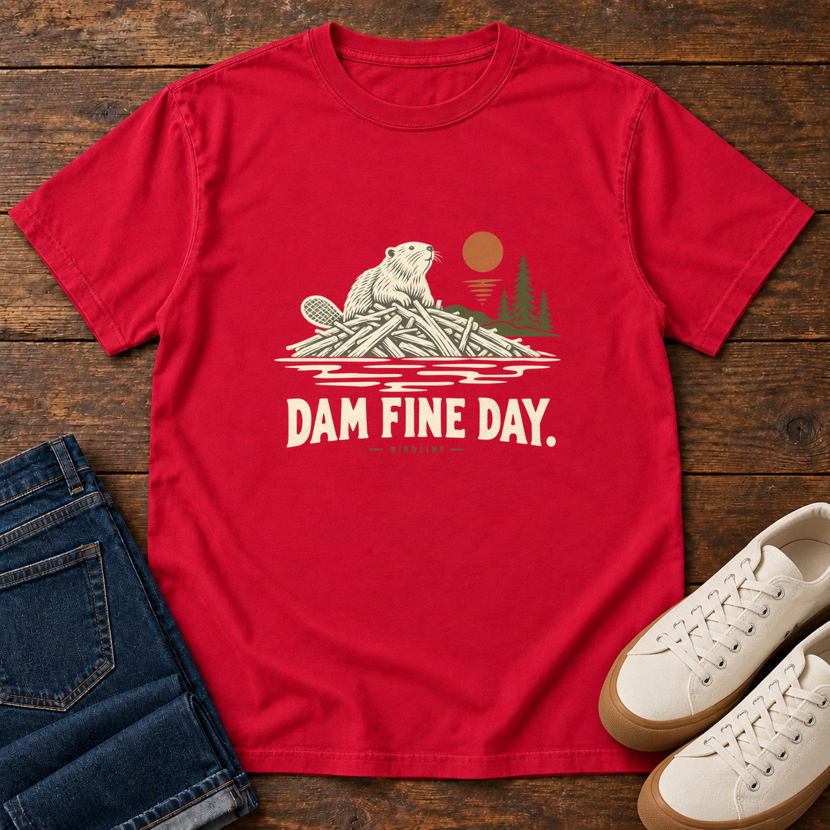 Red Dam Fine Day T-Shirt with a beaver on a pile of branches beside evergreens and an orange sun; front flat lay on wood.