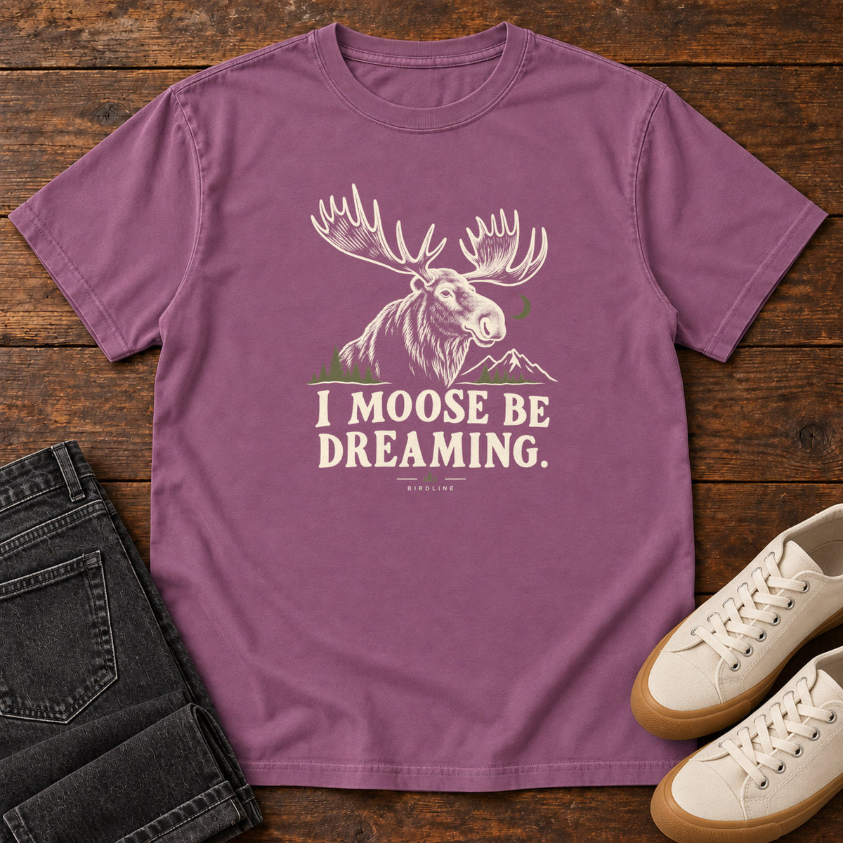 Berry I Moose Be Dreaming T-Shirt with a cream moose portrait with broad antlers, mountains and a crescent moon; front flat lay on wood.
