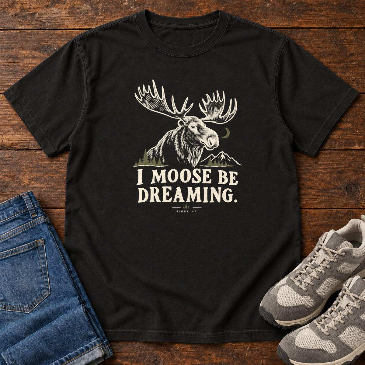 Black I Moose Be Dreaming T-Shirt with a cream moose portrait with broad antlers, mountains and a crescent moon; front flat lay on wood.