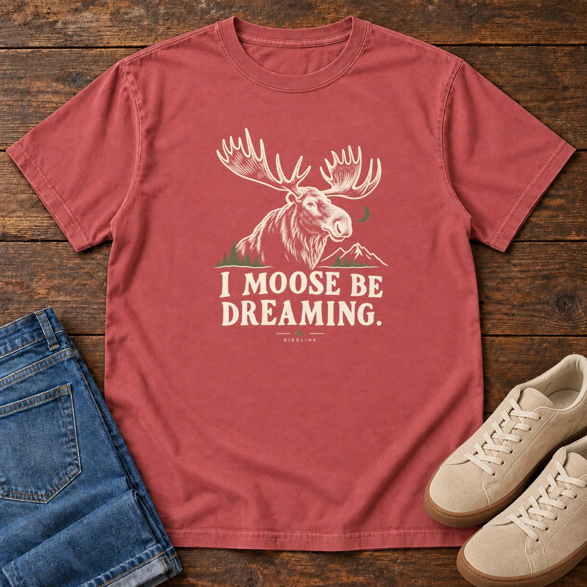 Crimson I Moose Be Dreaming T-Shirt with a cream moose portrait with broad antlers, mountains and a crescent moon; front flat lay on wood.