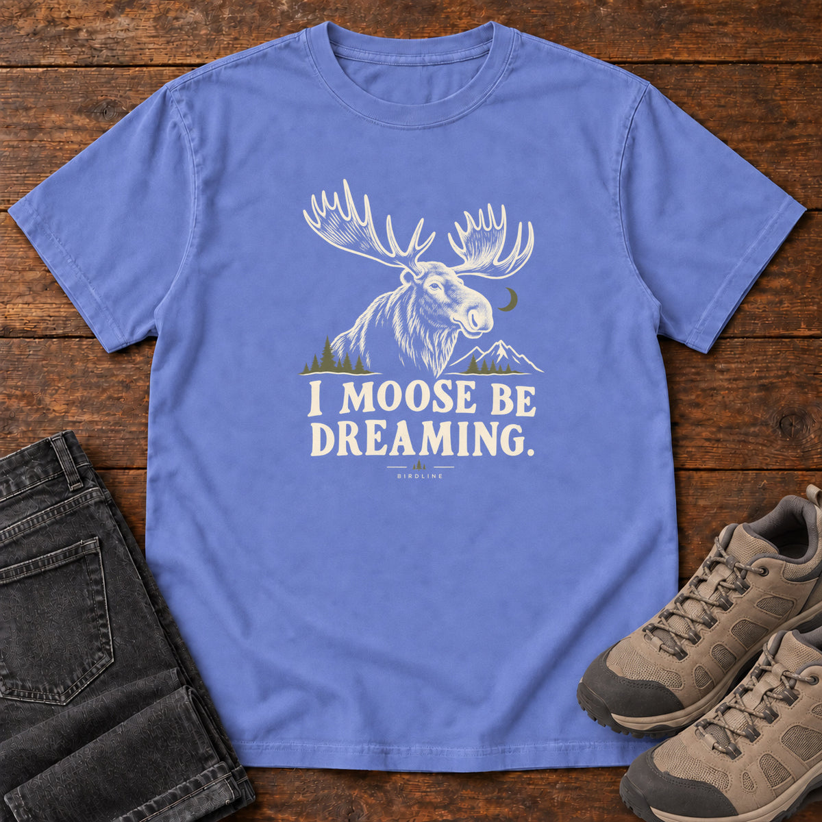 Flo Blue I Moose Be Dreaming T-Shirt with a cream moose portrait with broad antlers, mountains and a crescent moon; front flat lay on wood.