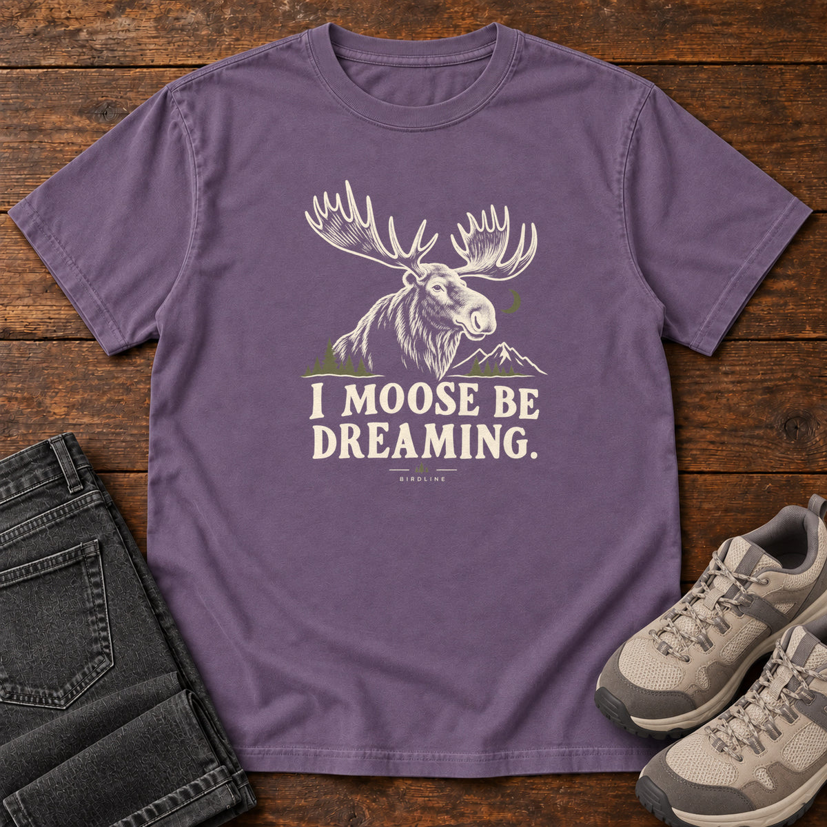 Grape I Moose Be Dreaming T-Shirt with a cream moose portrait with broad antlers, mountains and a crescent moon; front flat lay on wood.