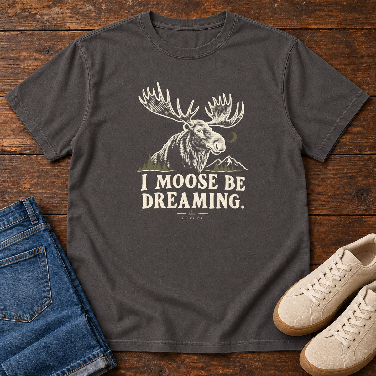 Graphite I Moose Be Dreaming T-Shirt with a cream moose portrait with broad antlers, mountains and a crescent moon; front flat lay on wood.