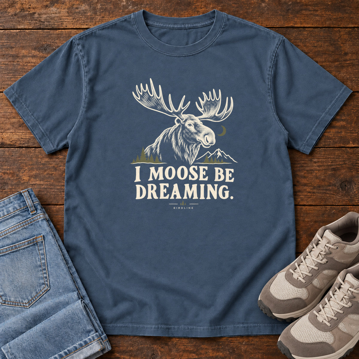 Midnight I Moose Be Dreaming T-Shirt with a cream moose portrait with broad antlers, mountains and a crescent moon; front flat lay on wood.